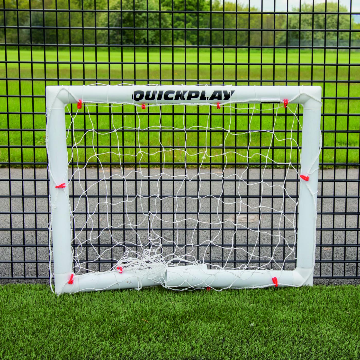 Q-FOLD Mini Football Goal 90 x 75 cm folded flat on artificial grass in front of a black mesh fence.