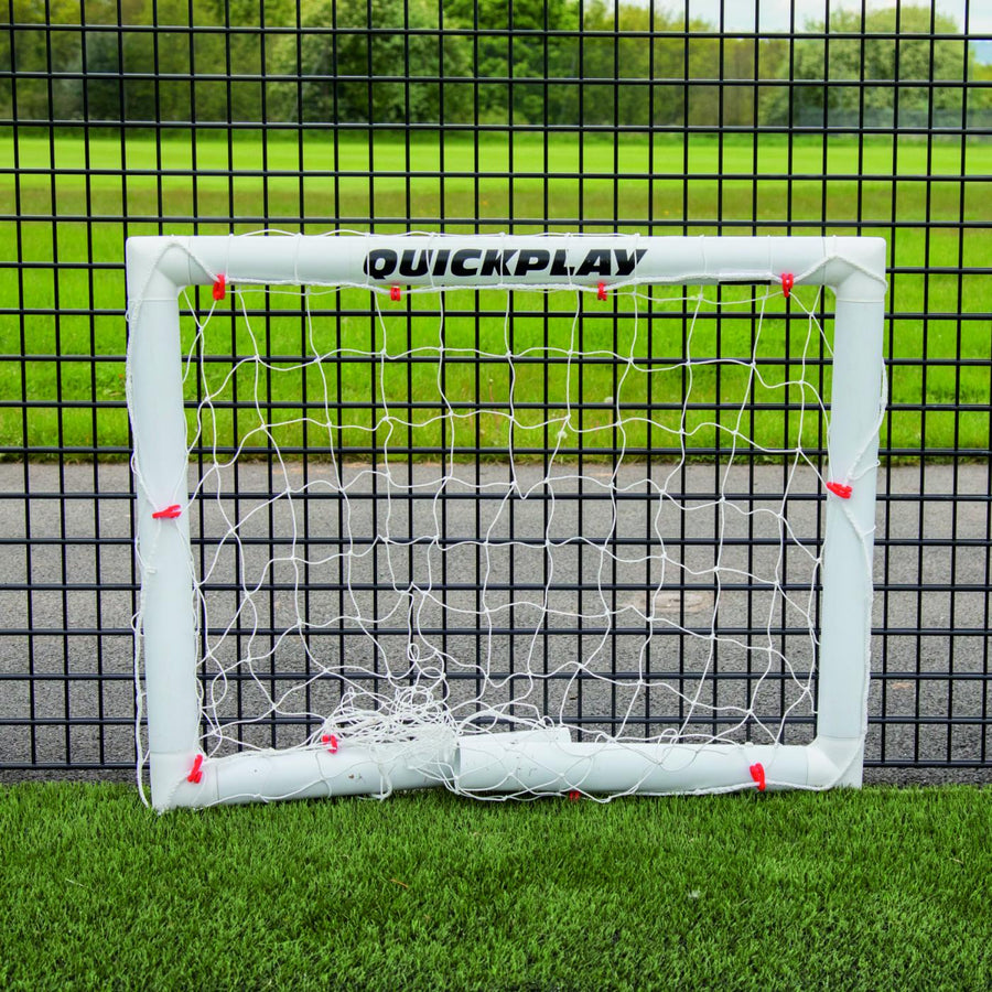 Q-FOLD Mini Football Goal 90 x 75 cm folded flat on artificial grass in front of a black mesh fence.