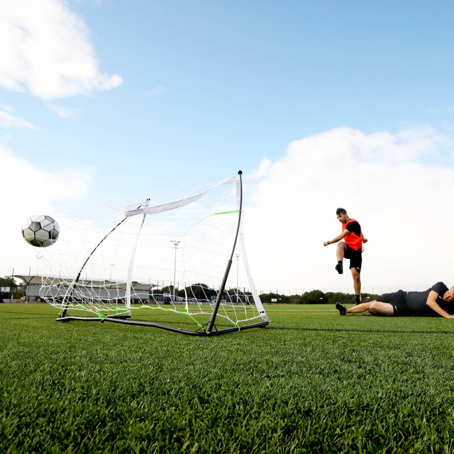 Low view of a player shooting a ball past the KICKSTER Elite 150 x 100 cm goal while another player lies on the pitch.