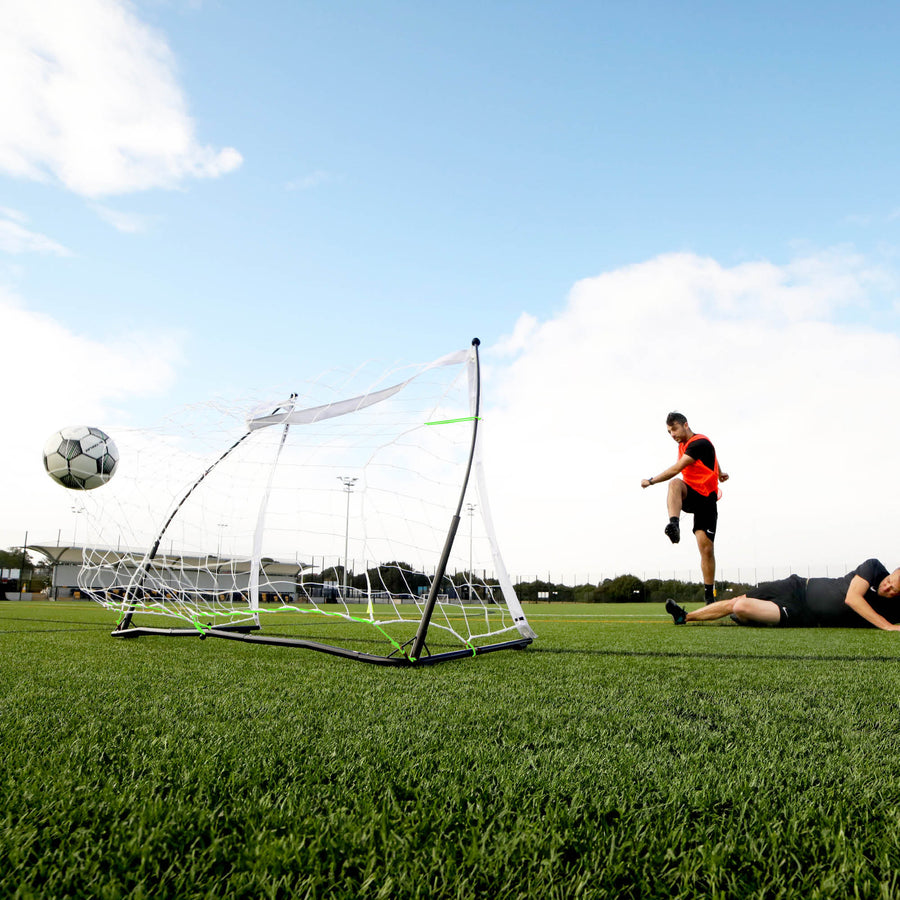 Low view of a player shooting a ball past the KICKSTER Elite 150 x 100 cm goal while another player lies on the pitch.