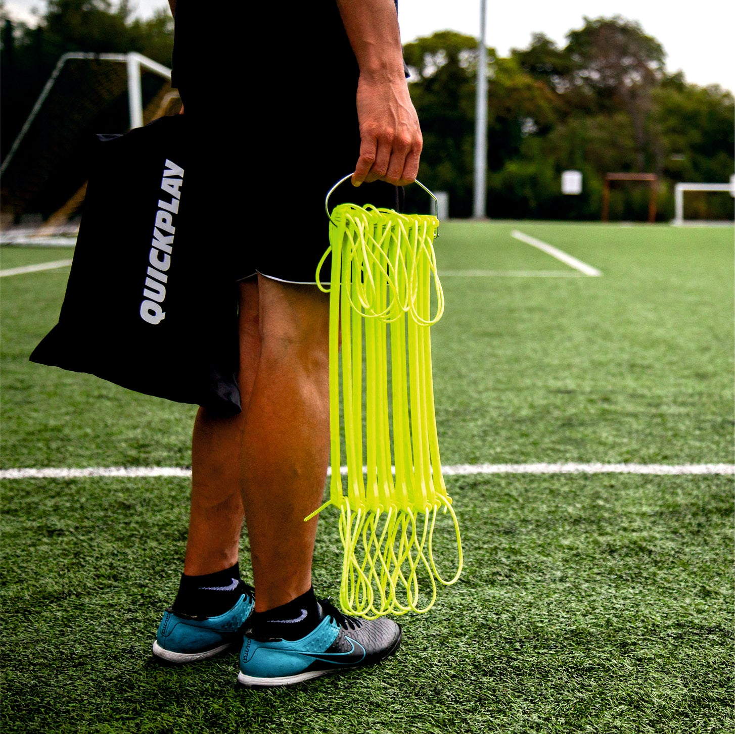 Player holding the folded yellow Agility Ladder on its hook, with a black QUICKPLAY bag, on an artificial pitch.