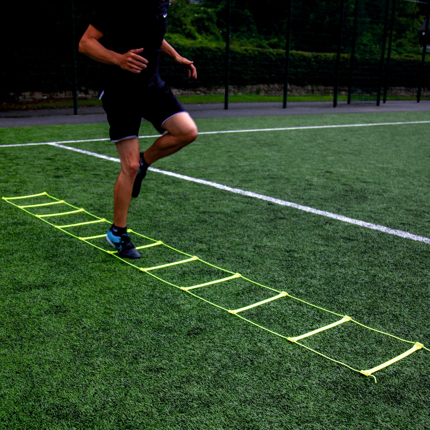 Player running through the yellow Agility Ladder on an artificial pitch, knees lifted.