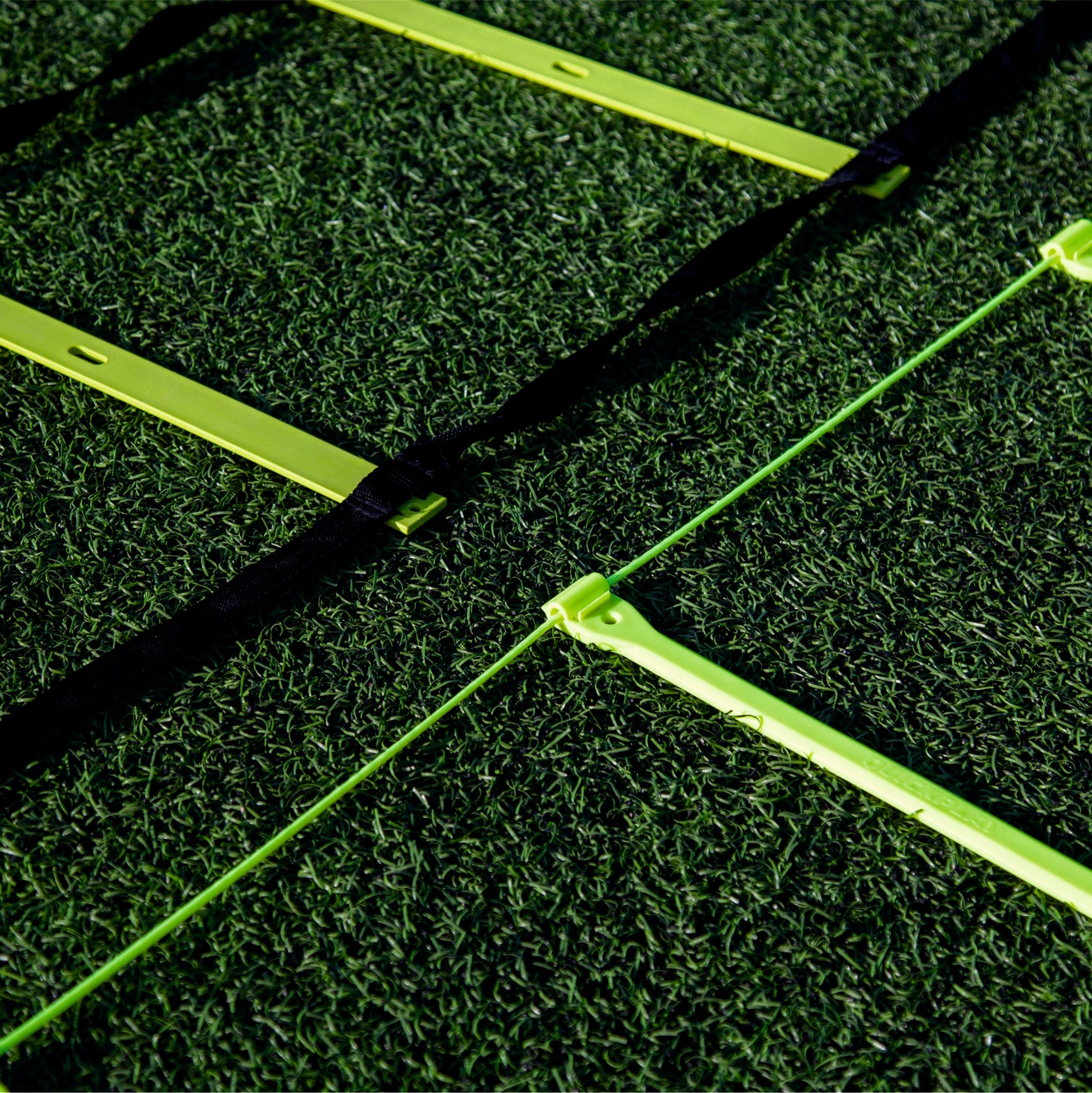 Close-up of yellow Agility Ladder rungs on artificial grass, one ladder with black straps and one with a thin green cord.