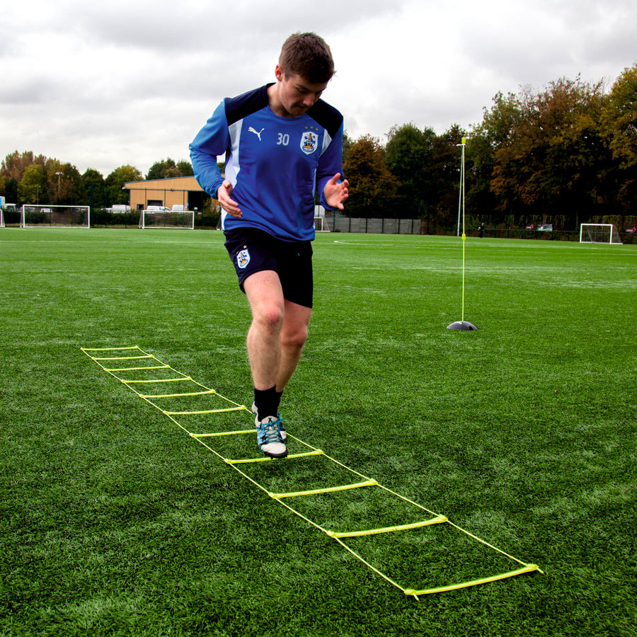 Player in a blue training top stepping through the Agility Ladder on an artificial pitch, with a pole behind.