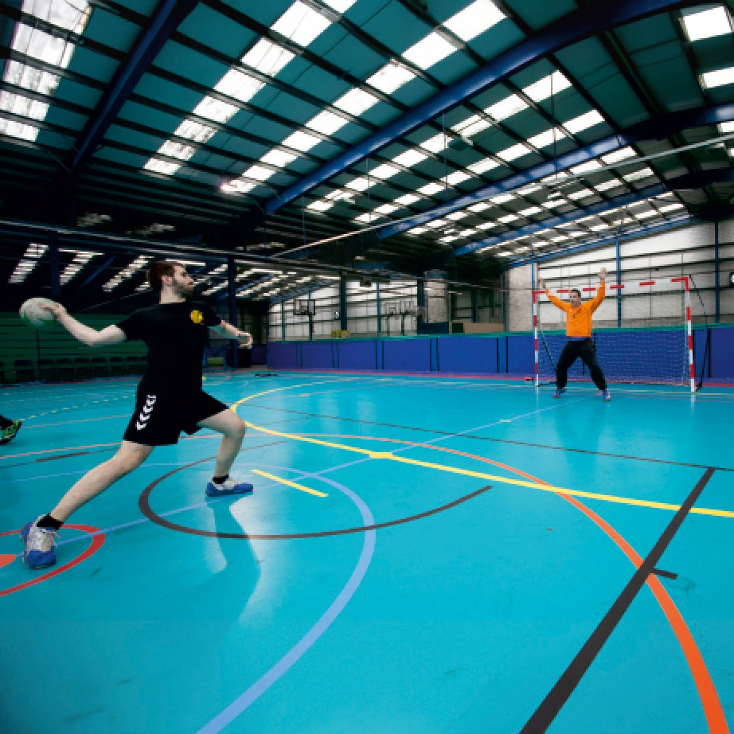 Player throwing a handball at a goalkeeper in front of the Portable Handball Goal Junior 240 x 170 cm in a large indoor hall.