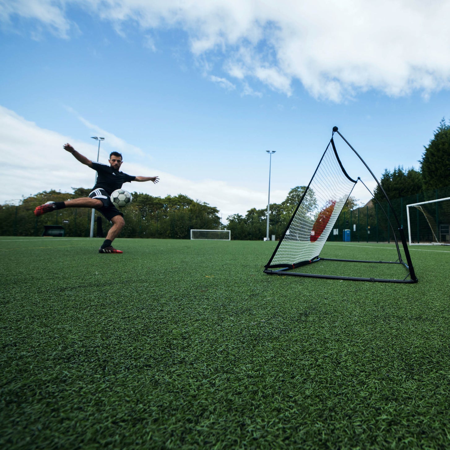 Player striking a volley towards the SPOT Football Rebounder 150 x 100 cm on an artificial pitch, seen from a low wide angle.