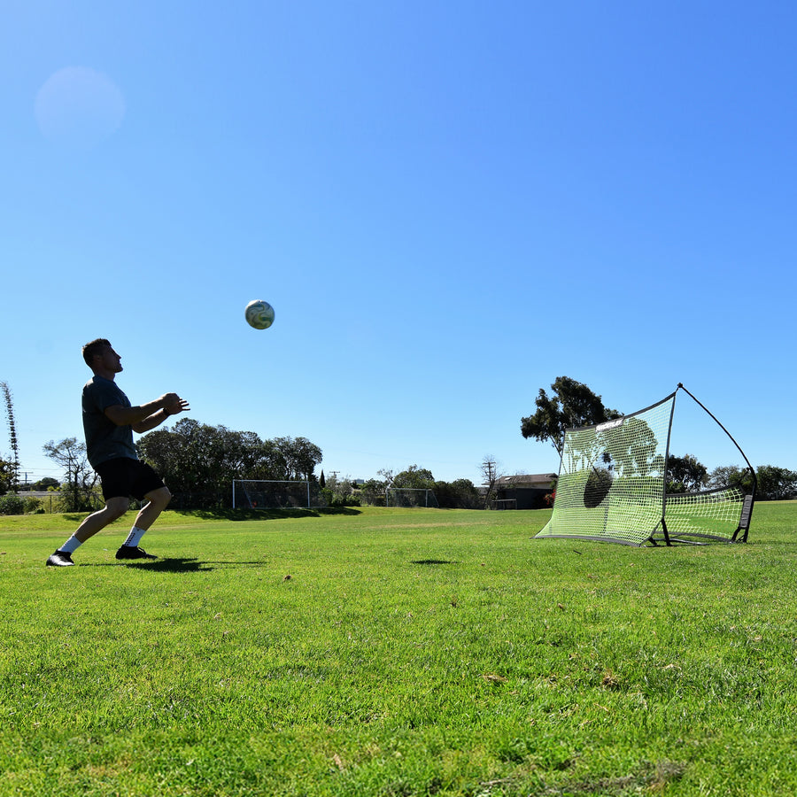 Wide view of a player preparing to control a ball in the air, with the TEKKERS Trainer rebounder behind him on grass.