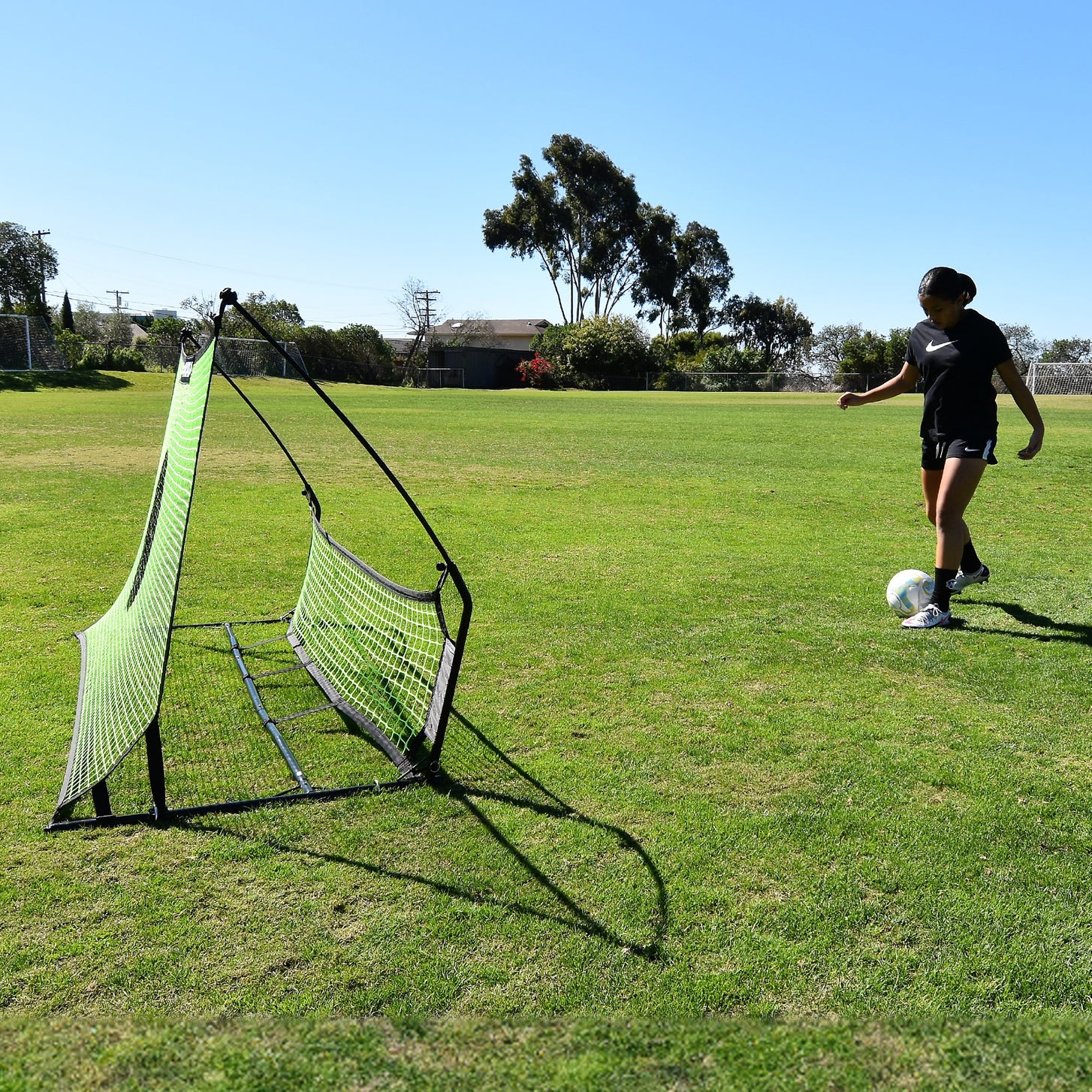 Player about to pass a ball into the TEKKERS Trainer Dual Net Football Rebounder, seen from the side on grass.