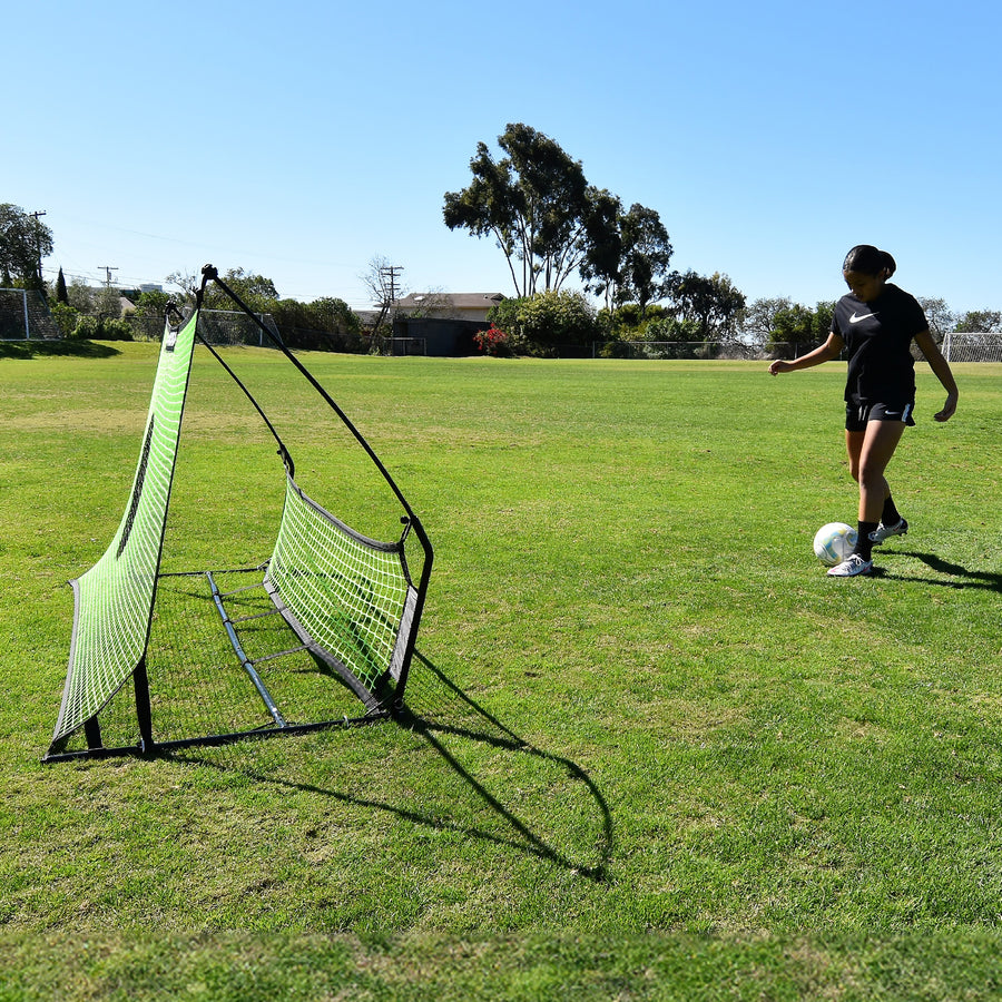 Player about to pass a ball into the TEKKERS Trainer Dual Net Football Rebounder, seen from the side on grass.