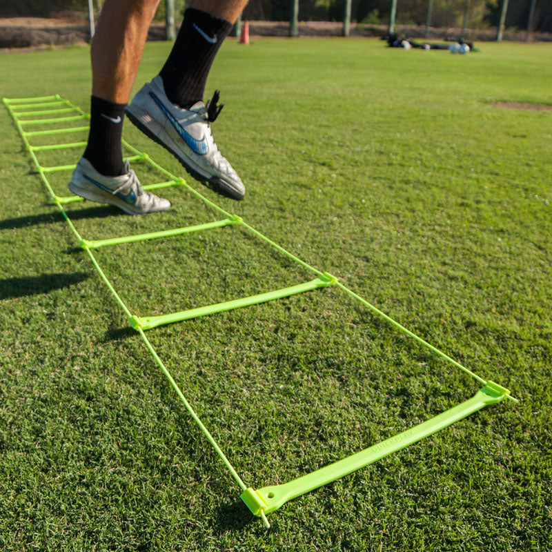 Player's feet stepping through the yellow Agility Ladder laid out on a grass pitch in sunshine.